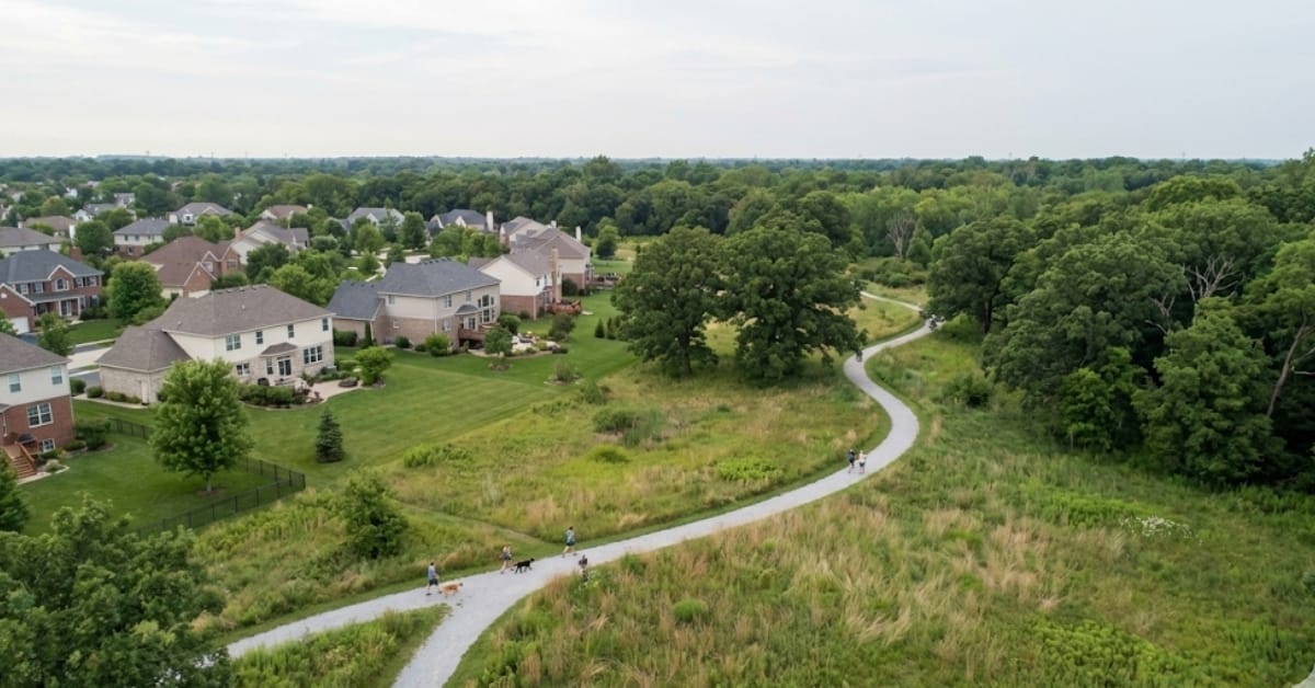 Aerial view of a residential neighborhood in Naperville bordering the Oldfield Oaks Forest Preserve, with a winding walking trail separating the homes from the dense woods.