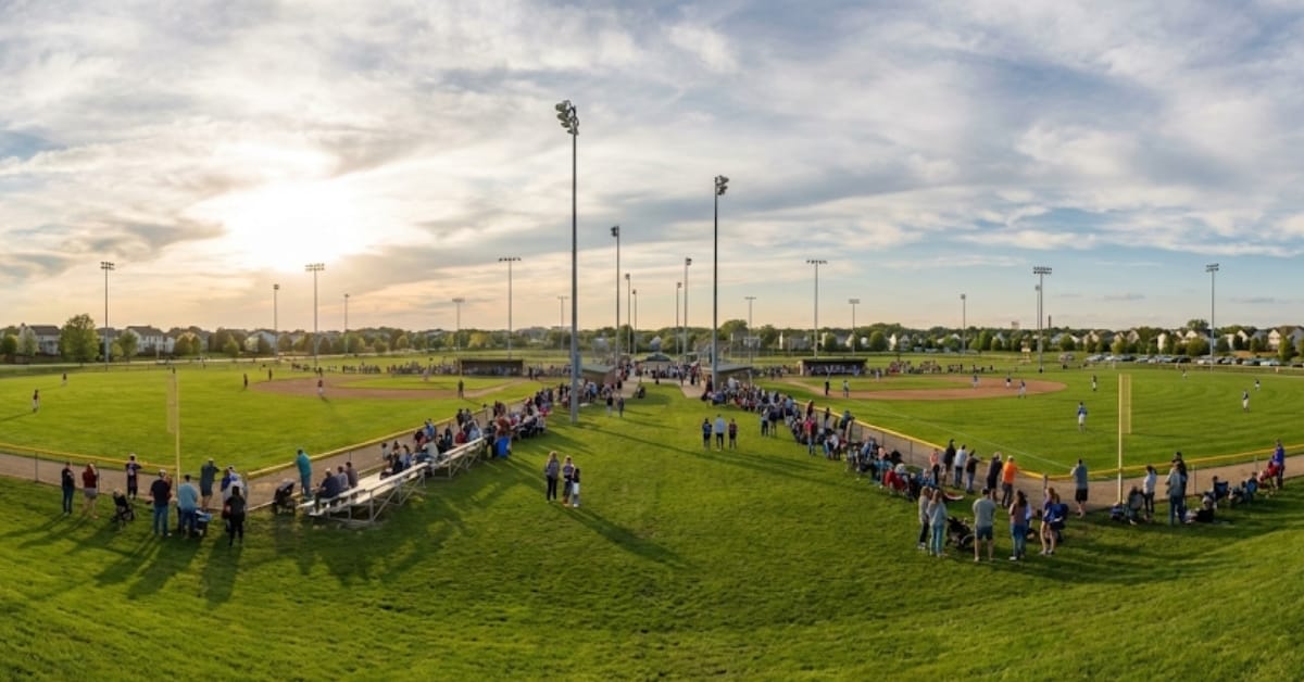 Panoramic view of the expansive Stuart Sports Complex in Aurora and Montgomery, featuring multiple baseball fields filled with players and spectators under stadium lights at sunset.