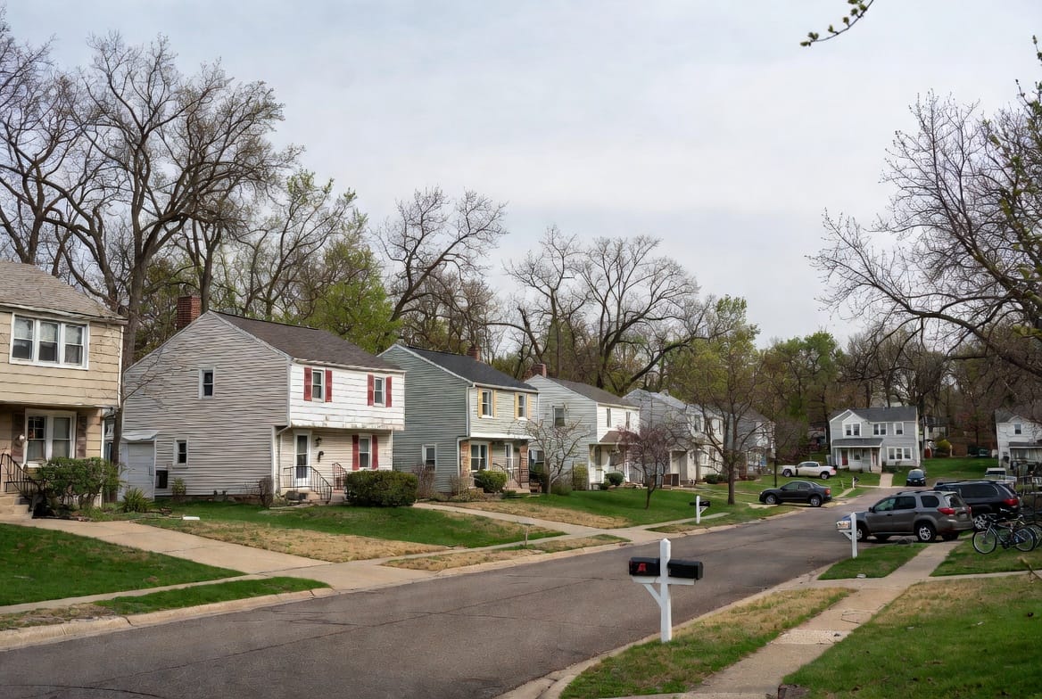 Naperville Real Estate Market 3 Quiet residential street in Naperville, Illinois, with a row of two-story suburban homes, small front lawns, leafing spring trees, parked cars, and mailboxes along the sidewalk under an overcast sky.