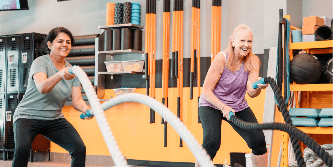 Two smiling women swing battle ropes in a personal training studio lined with orange racks of foam rollers and balls