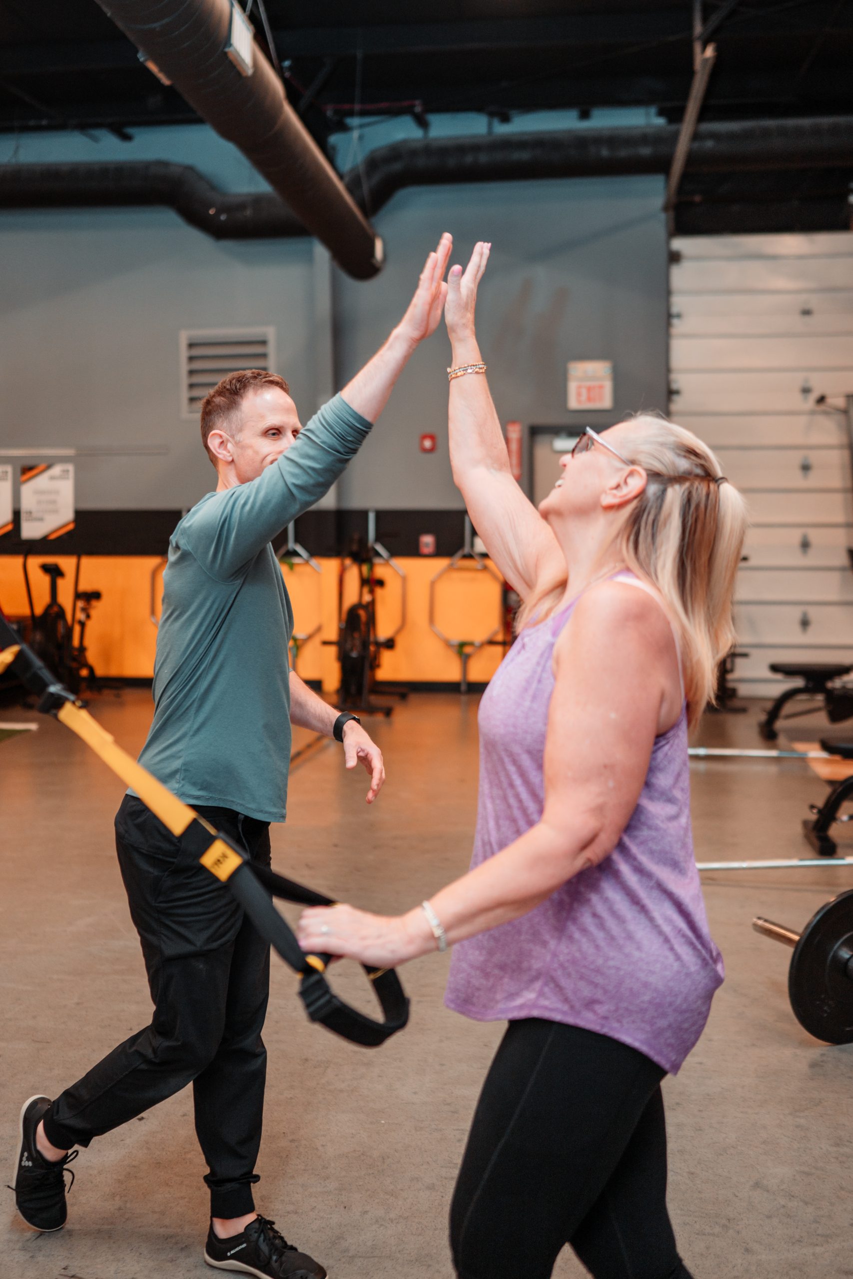 A coach in a teal shirt high fives a member holding a yellow suspension trainer strap inside a personal training studio
