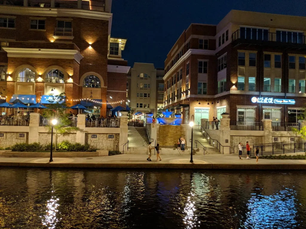 Night view of downtown Naperville Riverwalk with waterfront dining, illuminated buildings, and people walking along the river.