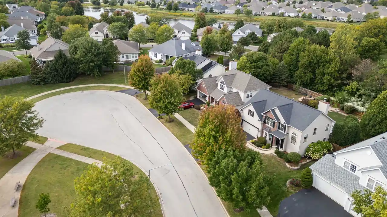 Aerial view of Heatherstone East subdivision in Naperville with residential homes, mature trees, sidewalks, and nearby pond.