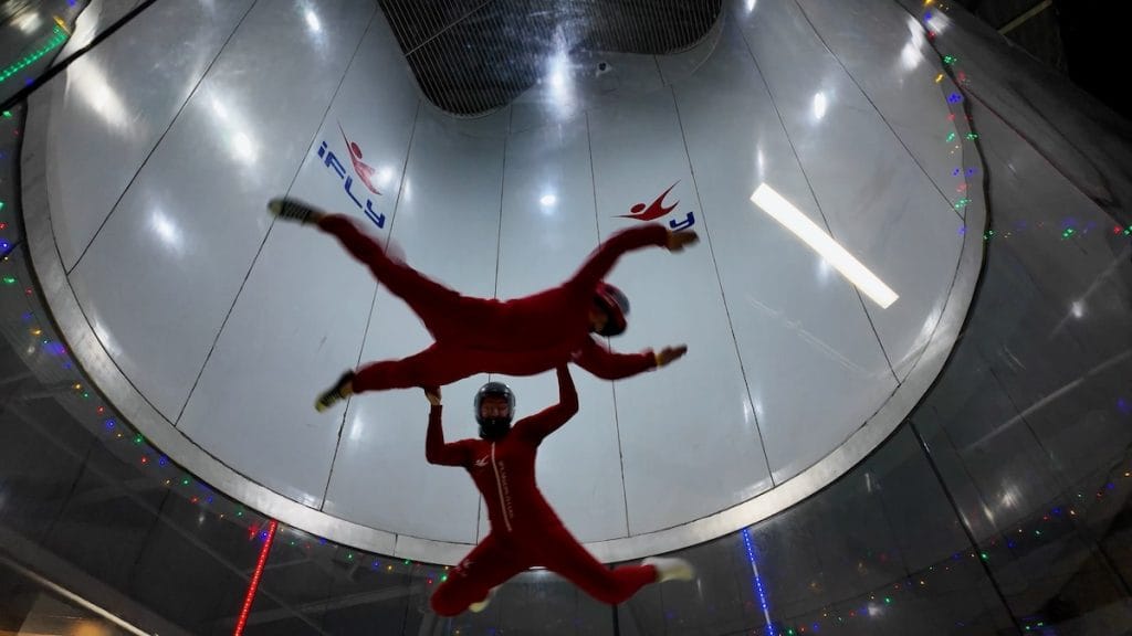 Overhead view of iFLY Naperville instructor in red flight suit guiding beginner flyer in vertical wind tunnel from below 2026