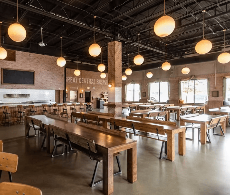 Brewery and dining space with long wooden tables, brick walls, pendant lighting, high ceilings, and bar area for architecture and interior design services in the Naperville area.