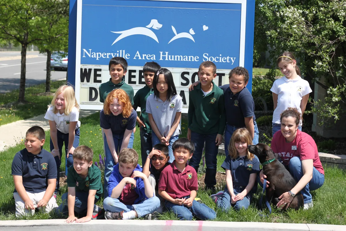 Children and staff gather with a dog in front of the Naperville Area Humane Society sign in Naperville, Illinois, highlighting a local nonprofit and family community visit
