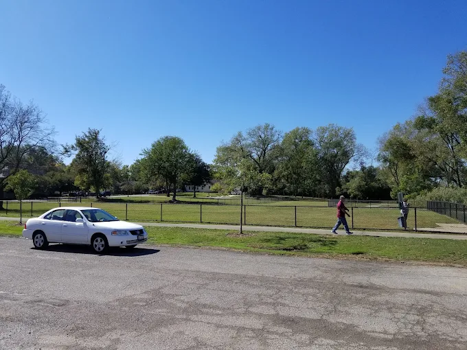 Fenced area at Phillips Park Dog Park in Aurora, Illinois with open grass, trees, sidewalk, and nearby parking.