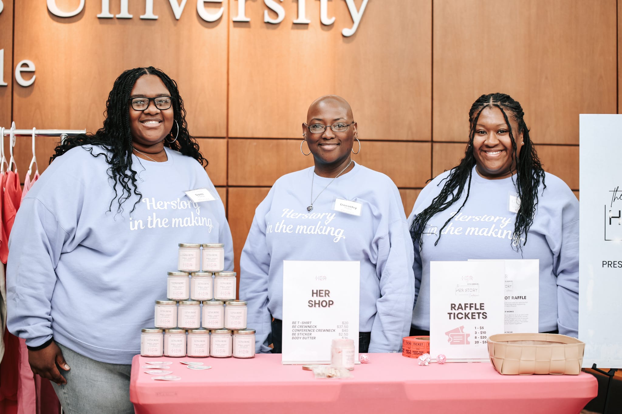 Beautifully EmpowHERed volunteers standing behind a fundraising and raffle table during a community event.