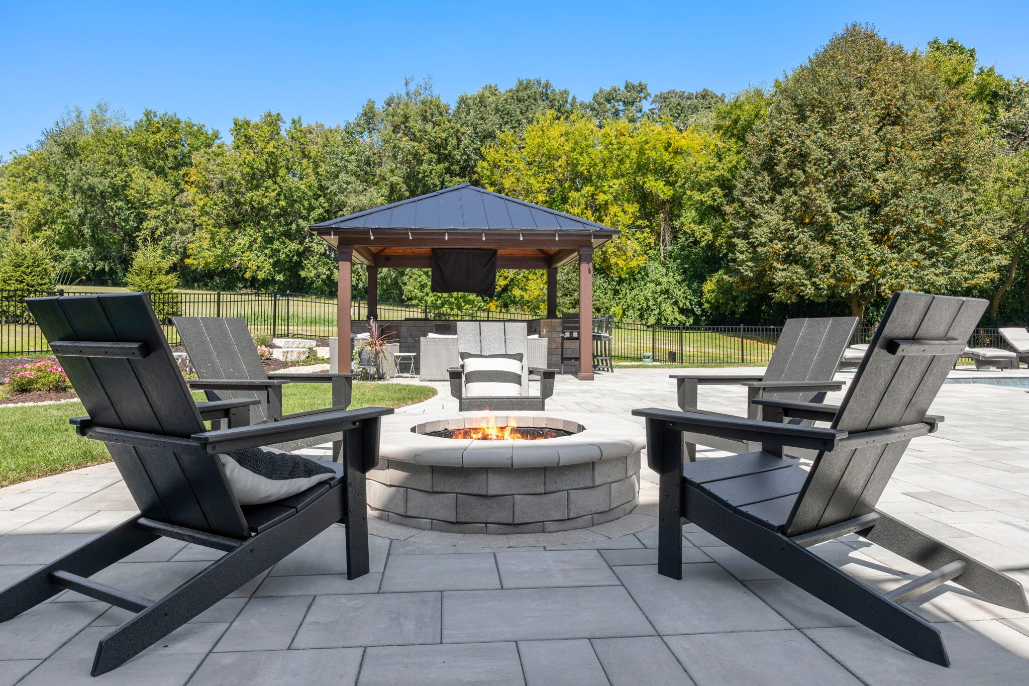 Black Adirondack chairs around a round paver gas fire pit with a covered pavilion and pool in the background
