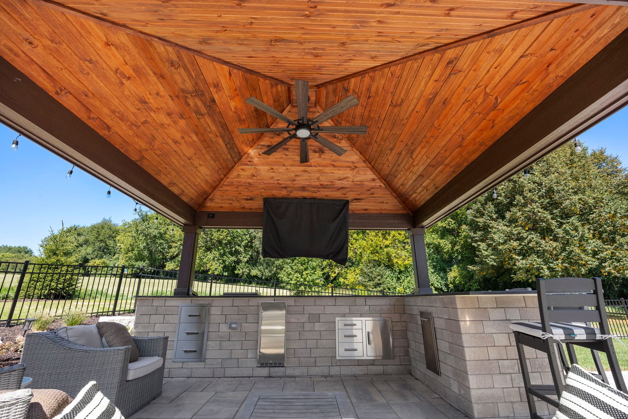 Covered pavilion interior with a tongue-and-groove wood ceiling, built-in grill, outdoor kitchen, and mounted TV