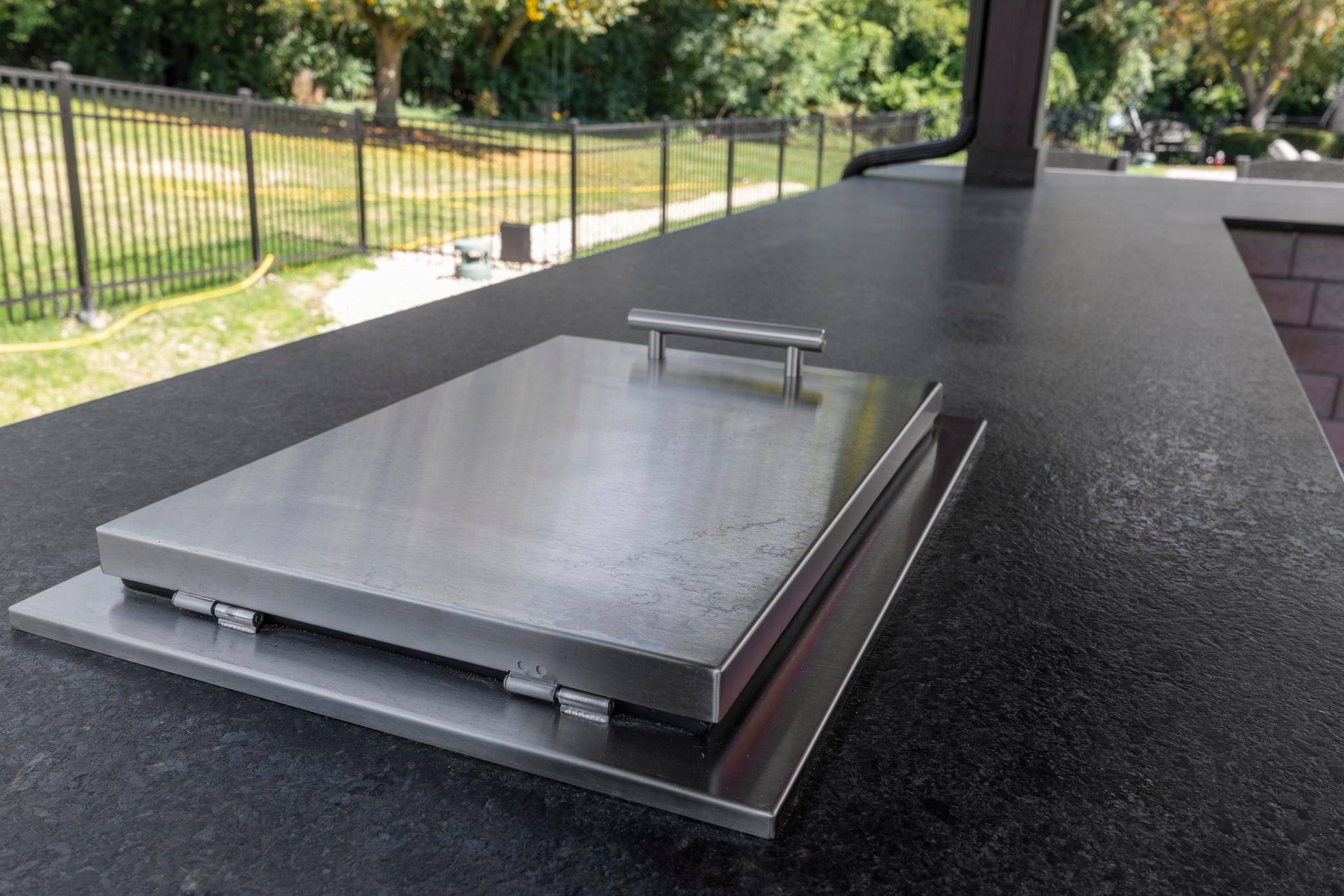 Close-up of a stainless steel built-in countertop insert with a hinged lid in a black granite outdoor kitchen