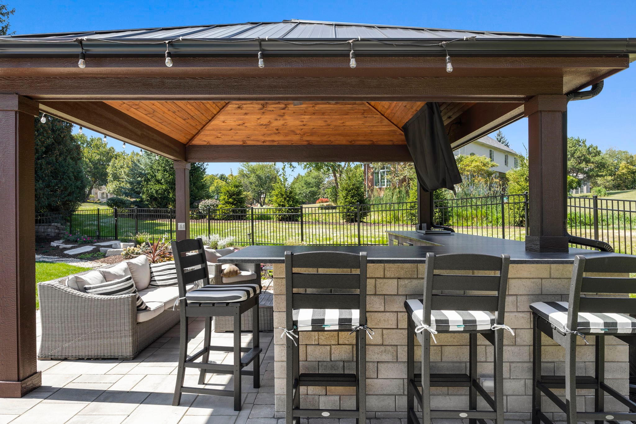 Covered outdoor bar with a stone base, granite top, and bar stools under a hardtop pavilion with string lights