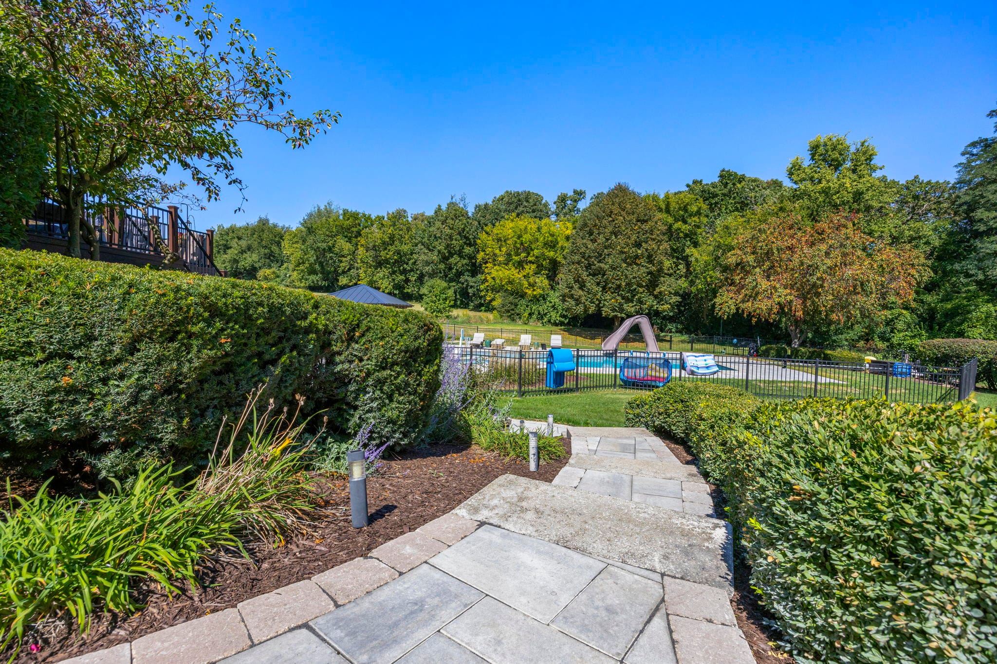 Paver walkway winding through trimmed hedges and planting beds toward a fenced backyard pool