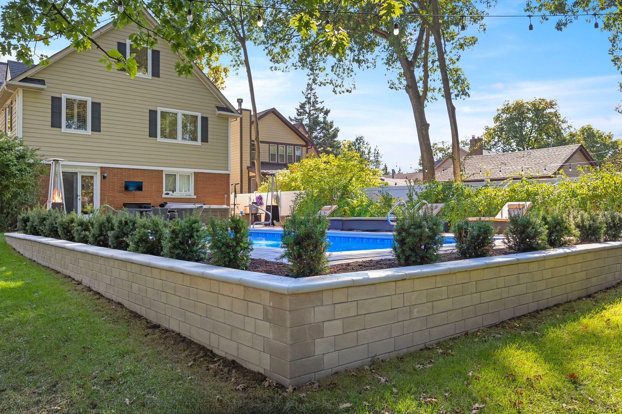 Tan modular block retaining wall enclosing a raised planting bed beside an in-ground pool with string lights