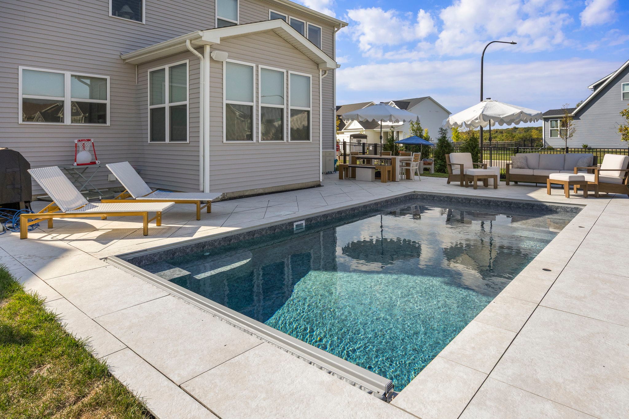 In-ground pool with a cream paver deck, chaise loungers, and a shaded dining area behind a two-story home