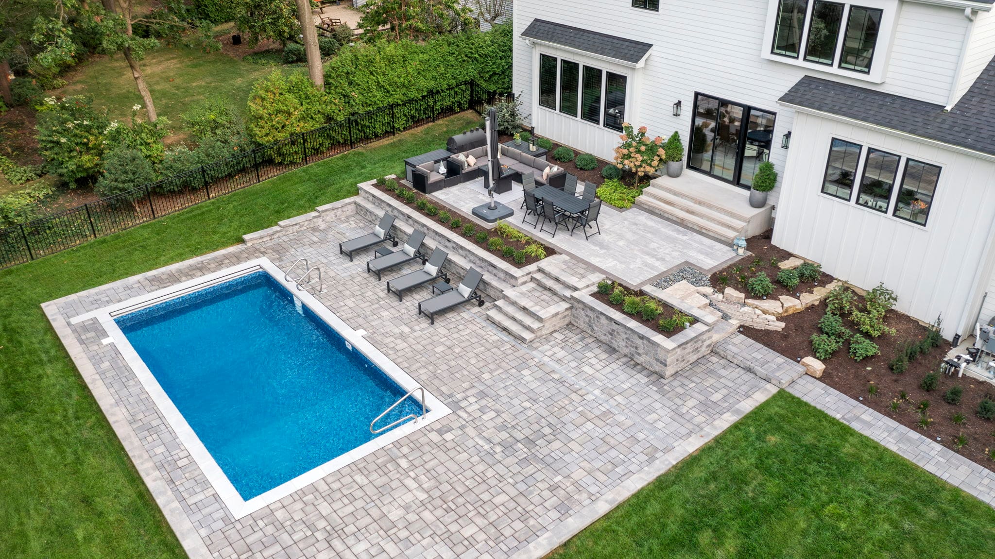 Aerial view of a rectangular in-ground pool and multi-level paver patio with outdoor dining behind a modern farmhouse