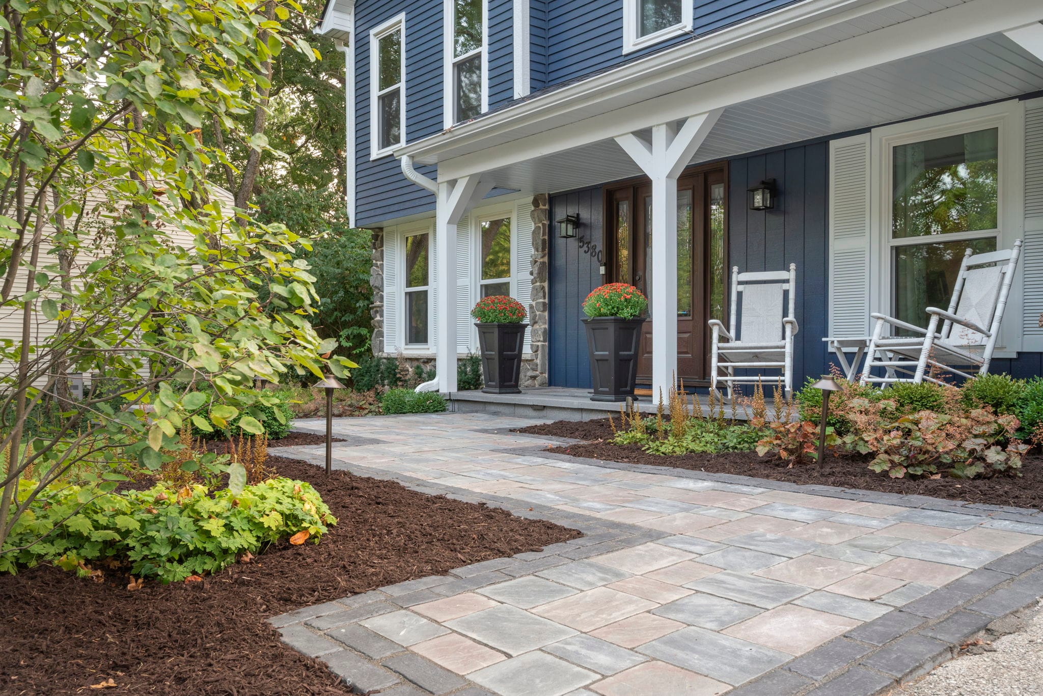 Paver front walkway with a stone border, landscape lighting, and planting beds leading to a navy blue home's porch
