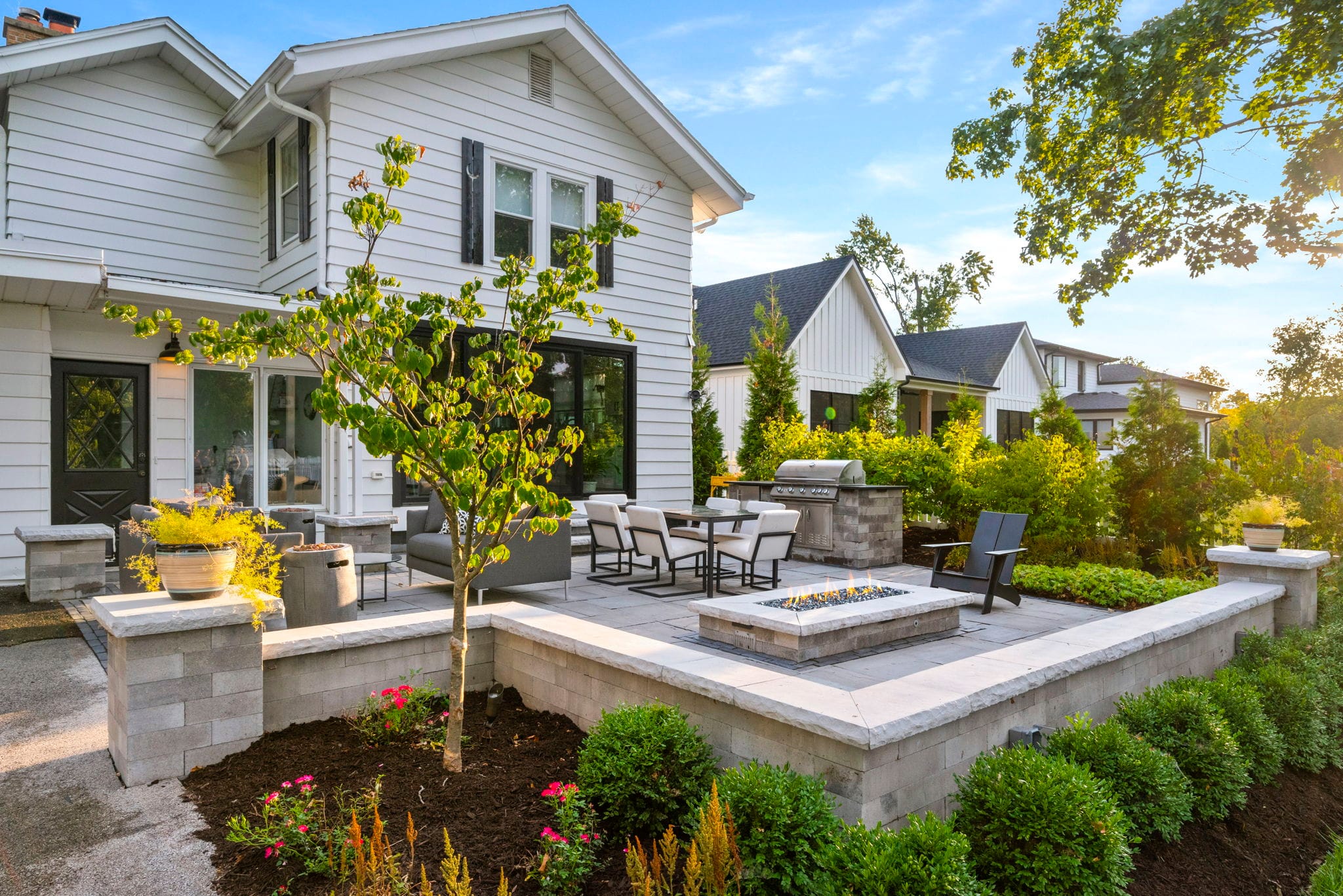 Raised paver patio with a seating wall, built-in grill, and gas fire pit behind a white farmhouse-style home