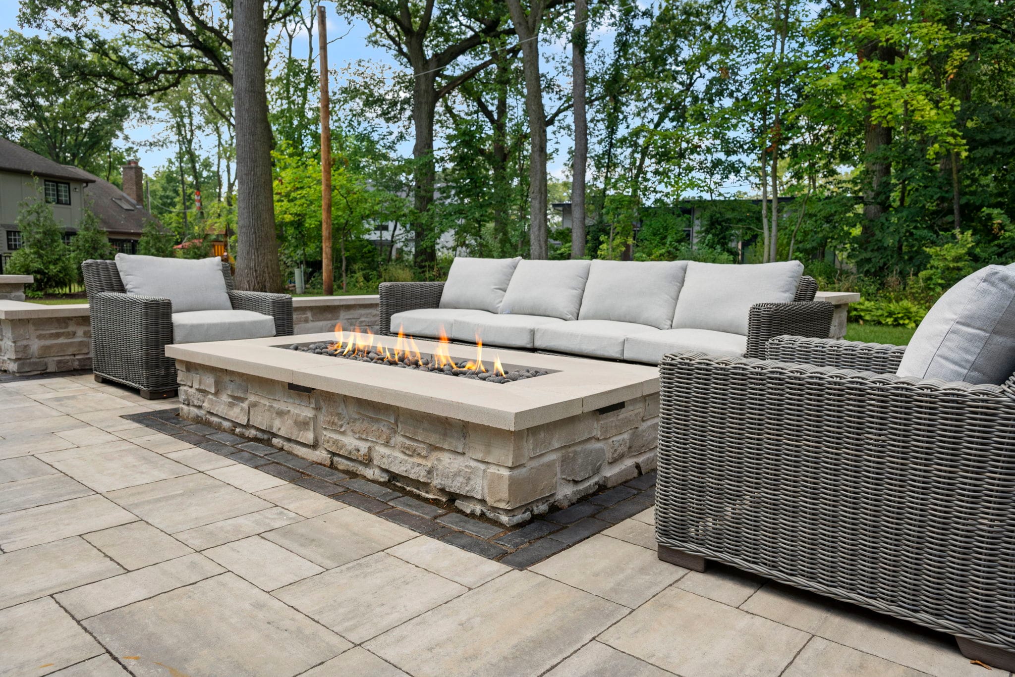 Long linear gas fire pit with a limestone base surrounded by gray wicker lounge furniture on a paver patio