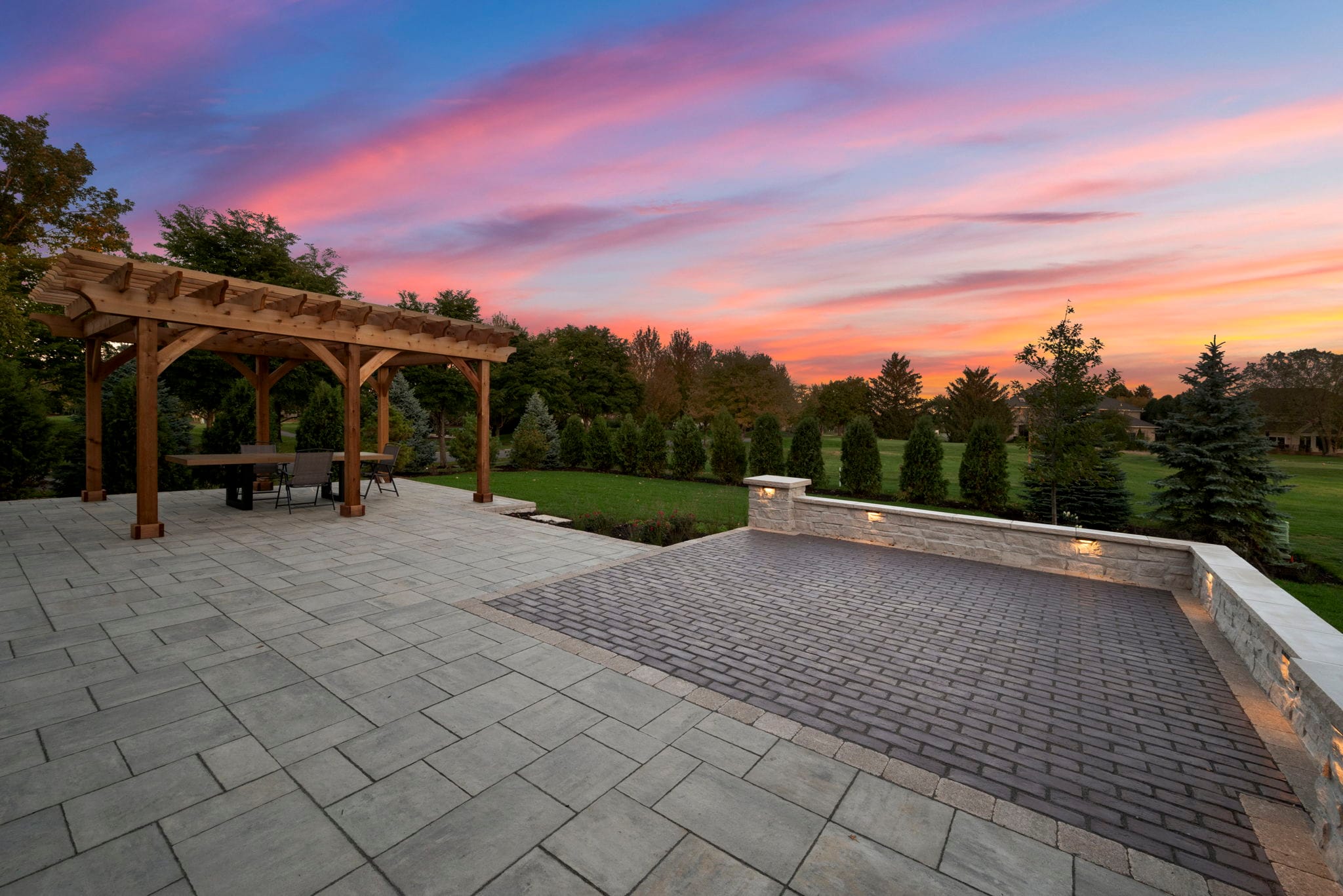 Paver patio at sunset with a wood pergola dining area and an illuminated seating wall with a contrasting paver inlay