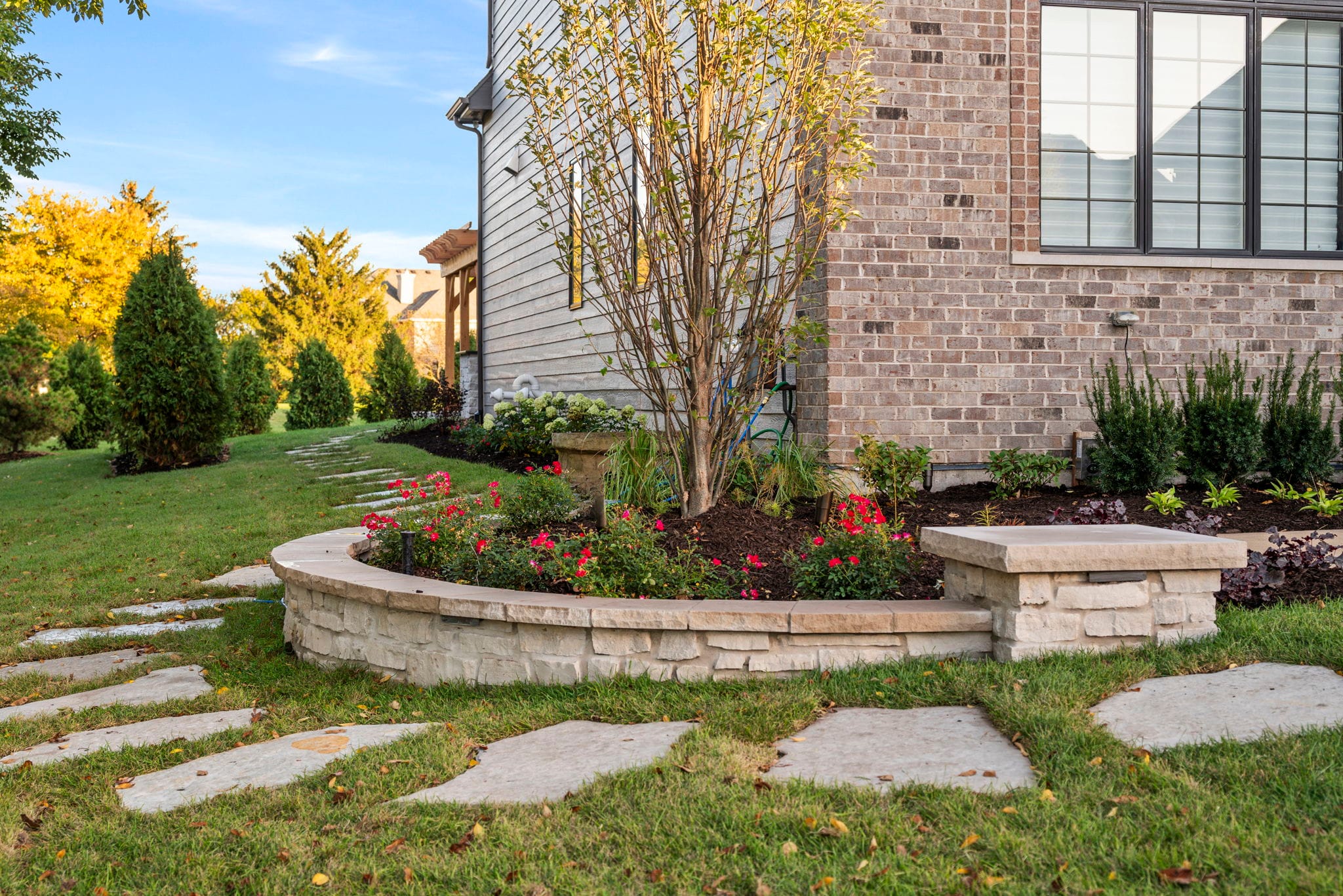 Curved stacked-stone seat wall and rose planting bed with a flagstone stepping-stone path beside a brick home