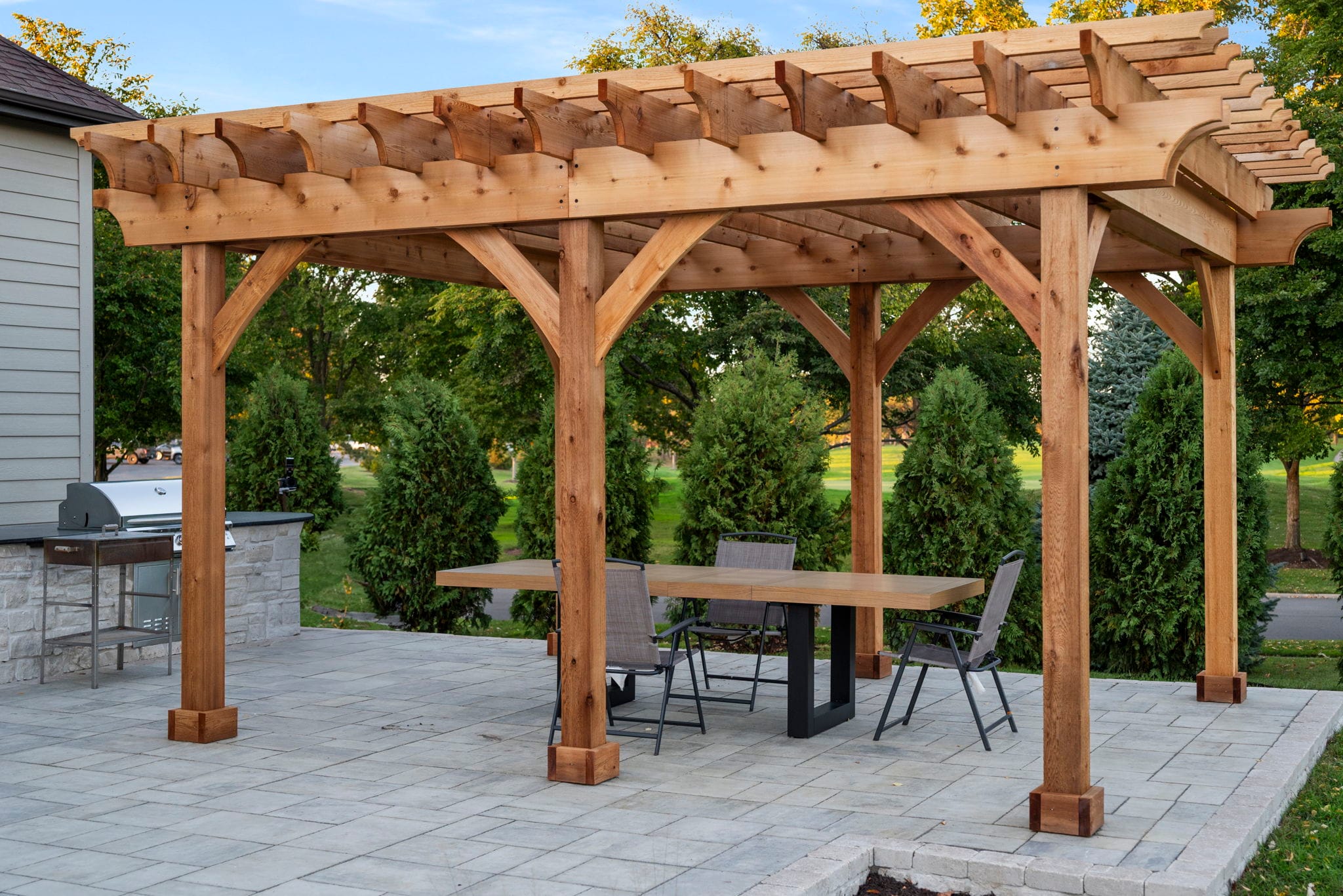 Cedar pergola over a paver patio dining area with a stone outdoor kitchen and arborvitae privacy screen