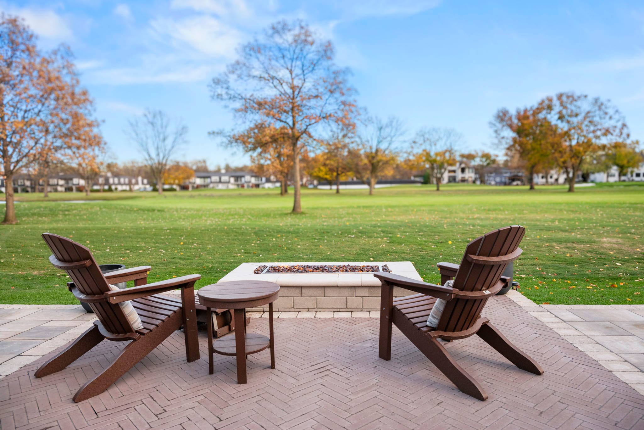 Two Adirondack chairs and a square gas fire pit on a herringbone brick paver patio overlooking open green space