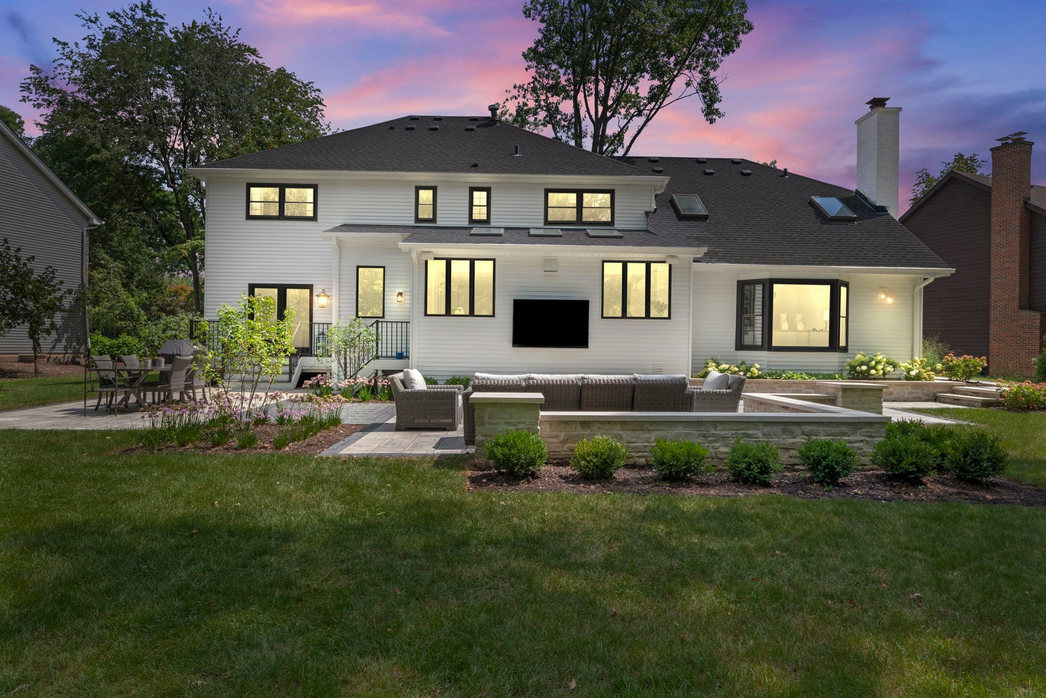 Twilight backyard of a white two-story home with a paver patio, seating wall, and outdoor lounge with a mounted TV