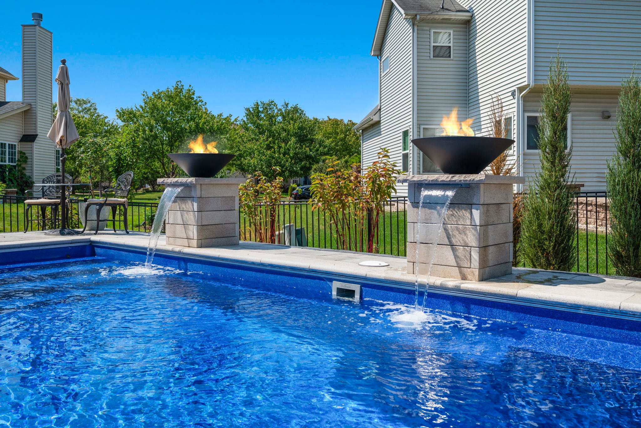 Two black fire-and-water bowls on stone pillars pouring into a blue in-ground pool on a sunny day